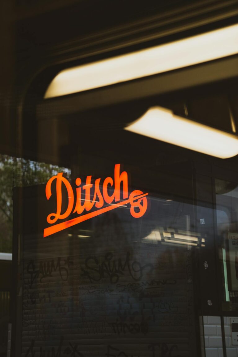 Reflection of orange neon Ditsch sign on a glass window with a blurred urban backdrop.
